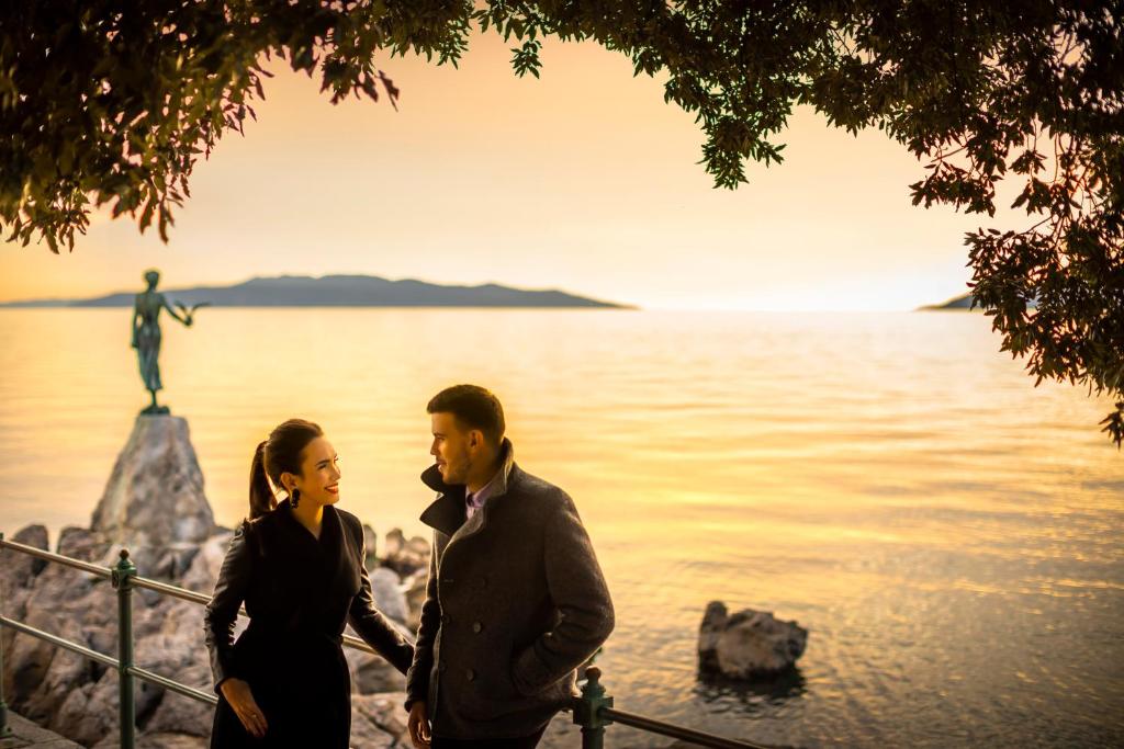 Romantiska ainava ar pāri, kas sarunājas pie jūras saulrietā, fons ar skulptūru un kalnainu ainavu.