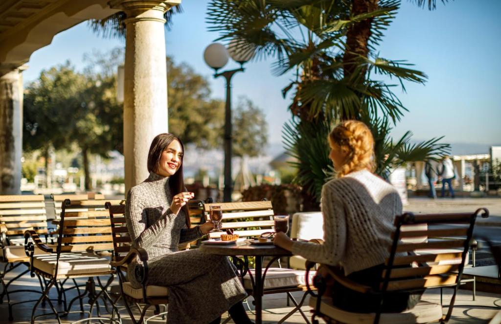 Divas sievietes sēž kafejnīcā zem palmām, baudot dzērienus un sarunājoties saulainā dienā. Gaisotne ir relaksējoša un draudzīga.