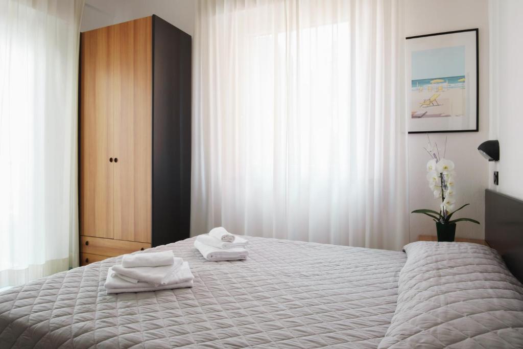 Mūsdienīga guļamistaba ar gultu, mīkstu segumu un baltām dvieļu kaudzēm, blakus liels koka skapis un ziedu kompozīcija uz naktsgaldiņa. Logi ar gaisīgām aizkariem nodrošina dabīgu apgaismojumu.