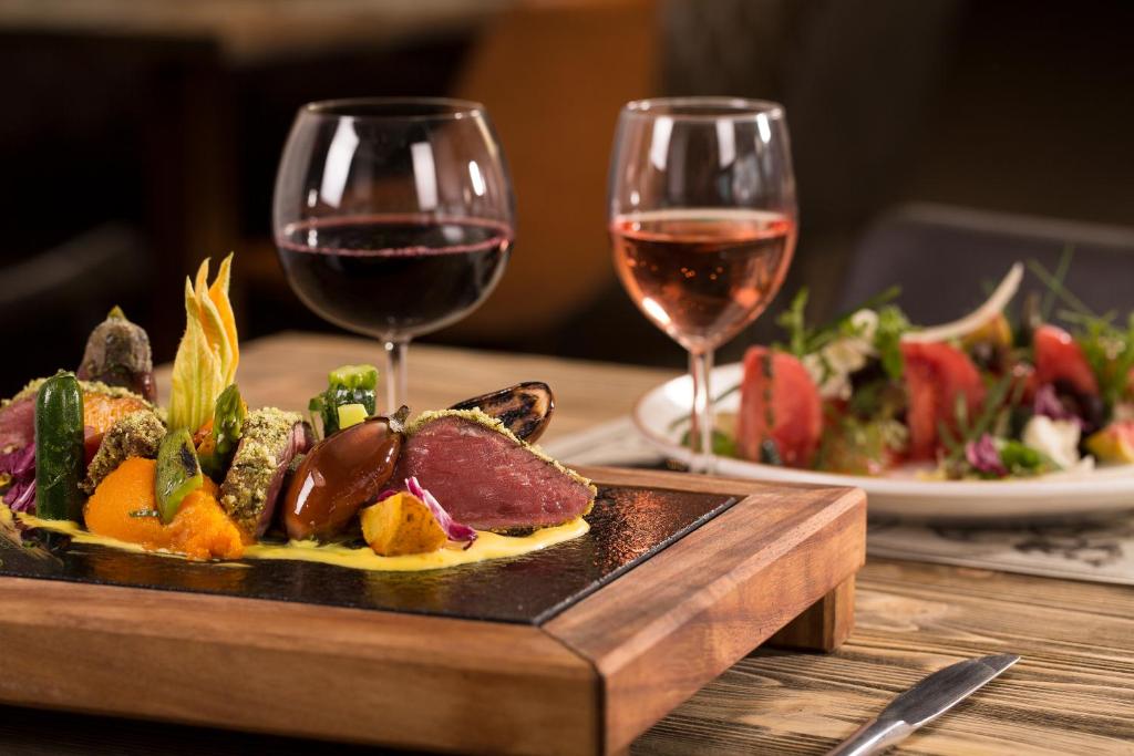 Gourmet ēdiens uz karstas akmens plāksnes ar dārzeņiem un mērci, blakus sarkanais un rozā vīns. Fonā redzama svaiga salātu porcija uz baltas šķīvja.