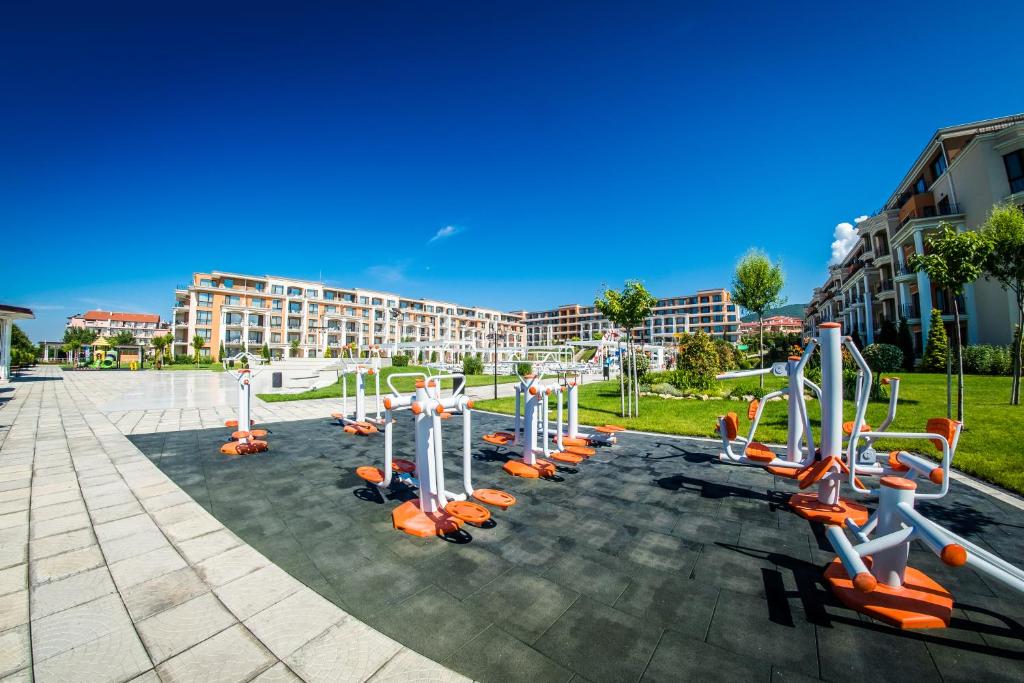 Ārtējo trenažieru zona ar modernām fitnesa iekārtām, kas ierīkota zaļā parkā, blakus dzīvojamām ēkām un skaidrām debesīm. Ideāla vieta aktīvai atpūtai un treniņiem.