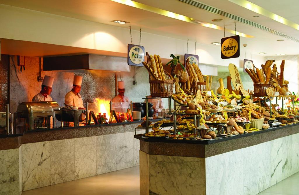 Bakerijas stends ar dažādiem konditorejas izstrādājumiem un maizēm, ko apkalpo šefpavāri. Gatavošanas zona ir redzama fonā, radot viesmīlīgu atmosfēru.