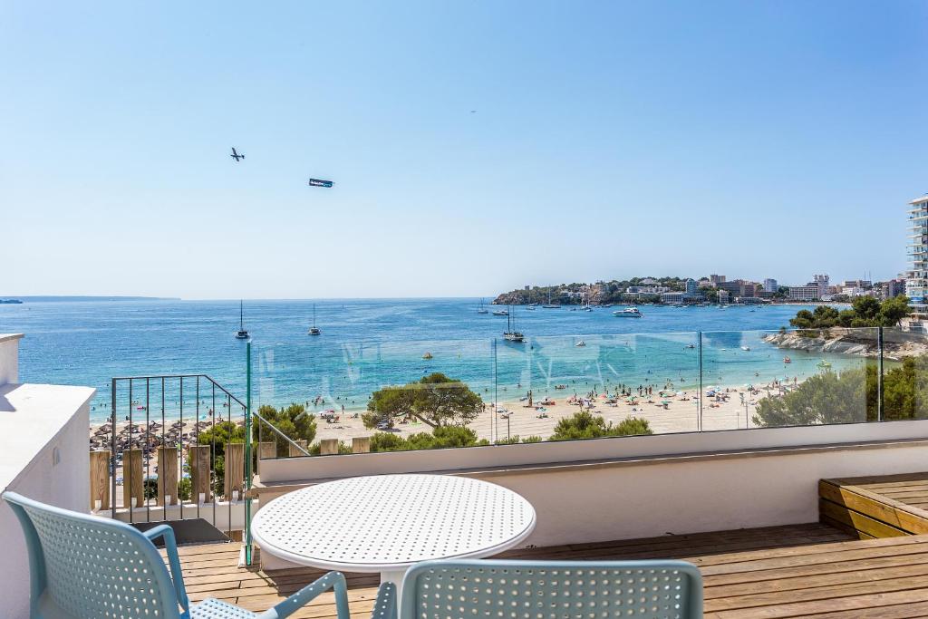 Jūras krasta skats ar pludmali un laivām, saulaina diena. Terase ar galdu un krēsliem, fonā redzami cilvēki, kas atpūšas pludmalē.