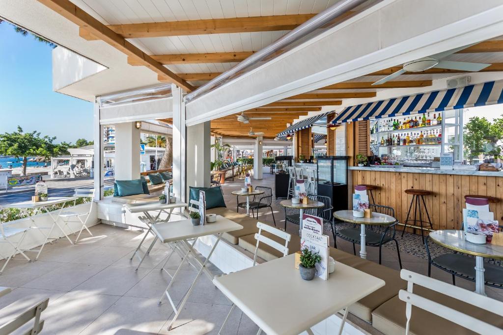 Moderns kafejnīcas interjers ar atvērtu terasi un skatu uz pludmali. Kafejnīcā ir eleganti galdi un krēsli, kas aicina apmeklētājus baudīt dzērienus un ēdienus.