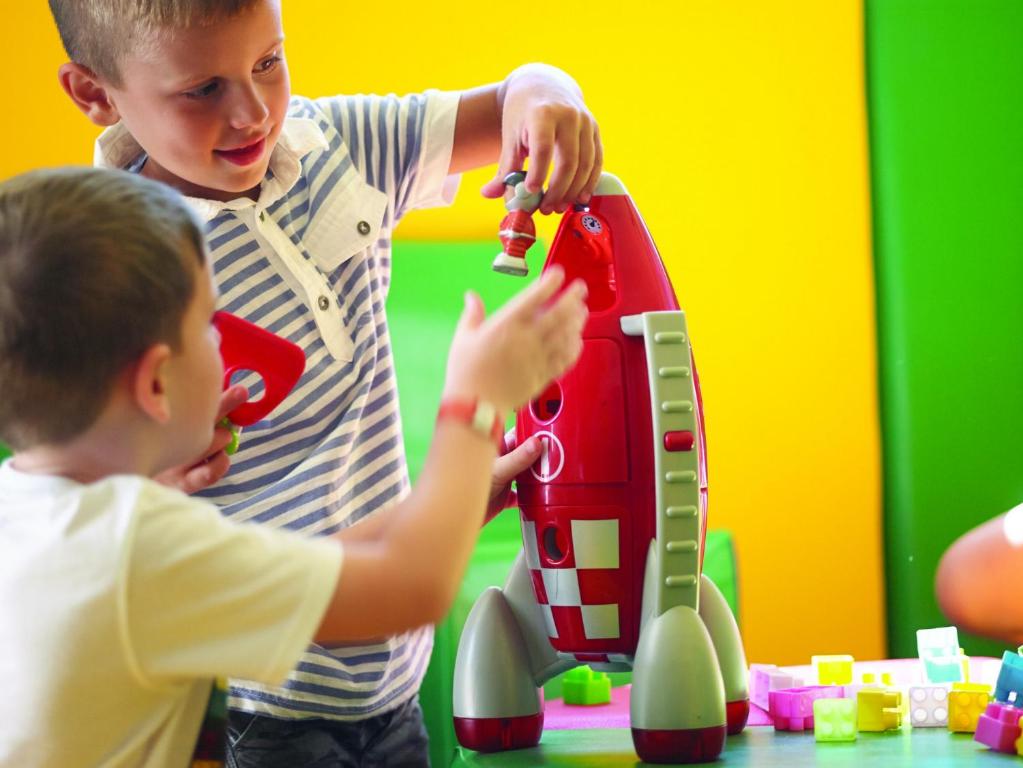 Divi bērni spēlējas ar sarkano raķeti, viens no viņiem pievieno figūru raķetes augšpusē, kamēr otrs tur sarkanu kontrolieru. Apkārt ir krāsaini rotaļlietu bloki.