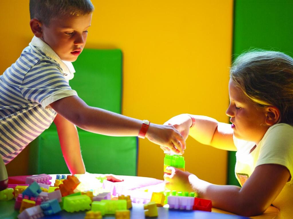 Bērni spēlējas ar krāsainām konstrukciju klucīšiem, būvējot torņus. Apgaismojums rada jautru un radošu atmosfēru.