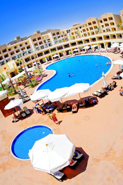 Liels baseins ar saules gultām un lietussargiem, ap kuru ir viesnīcas komplekss. Apkārt baseinam ir apmeklētāji, kas atpūšas un peldas.