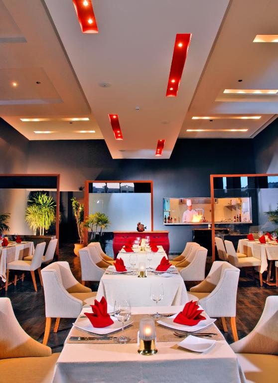 Mūsdienīgs restorāns ar eleganti klātiem galdiem, sarkanām salvetēm un stilīgu apgaismojumu. Interjerā redzami augi un atvērta virtuve, radot patīkamu atmosfēru.