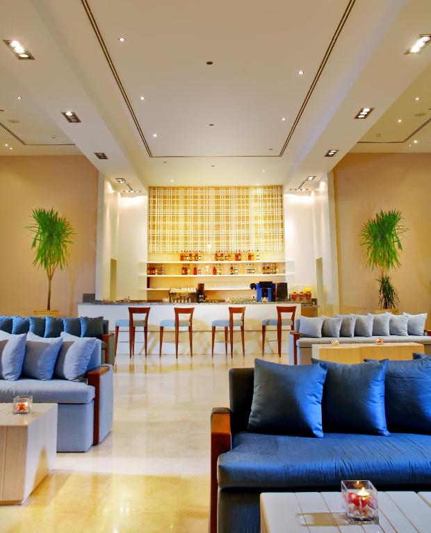 Mūsdienīgs viesnīcas bārs ar elegantiem dīvāniem un baristu zonu, kas rotā telpu ar zaļām augu kompozīcijām. Gaismas un interjera dizains rada patīkamu un relaksējošu atmosfēru.