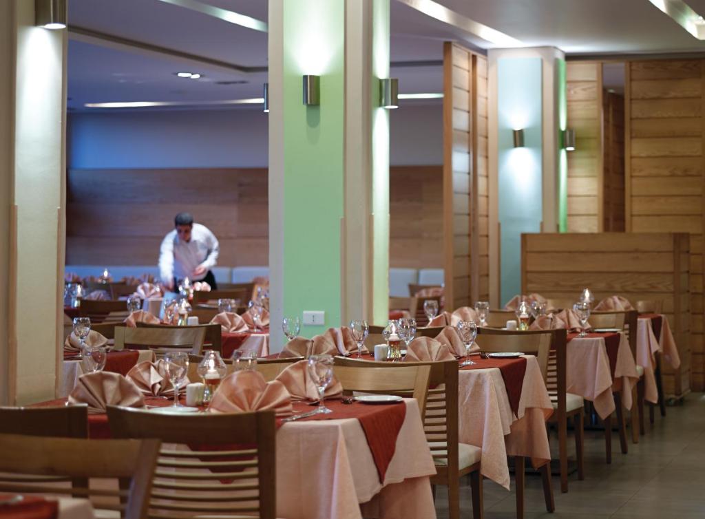 Elegants restorāna interjers ar galdiem, kas klāti ar rozā galdautiem un dekoratīvām salvetēm. Apkalpojošais darbinieks kārto galdu, radot patīkamu atmosfēru.