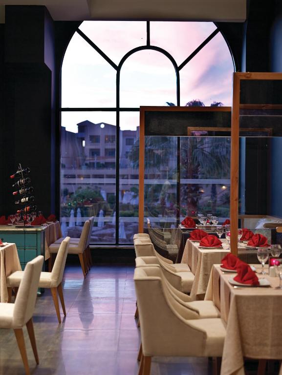 Elegants restorāna interjers ar mēbelēm un galdiem, kas klāti ar sarkaniem salvetēm, un liela loga ēka, caur kuru redzams saulriets. Mūsdienīgs dizains un patīkama atmosfēra.