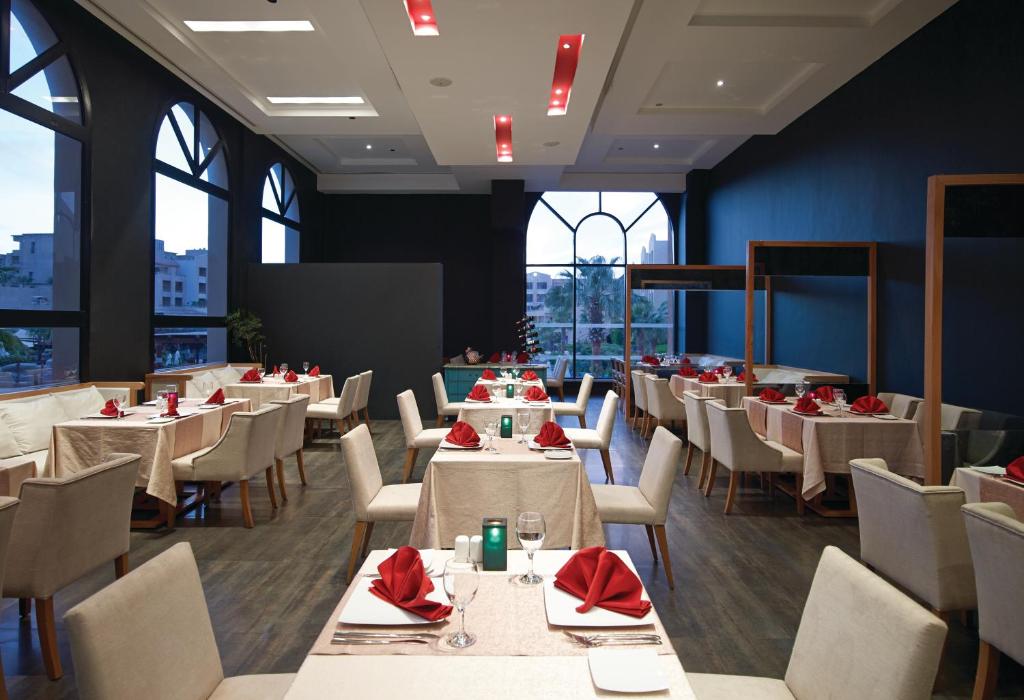 Mūsdienīgs restorāna interjers ar elegantiem galdiem, kas klāti ar baltiem galdautiem un sarkaniem salvetēm. Lielie logi piedāvā skatu uz ārpusi, radot patīkamu atmosfēru.