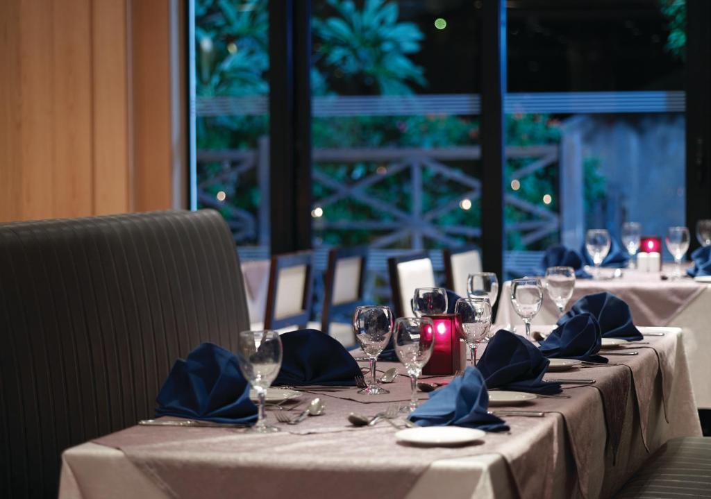 Elegants restorāna iekārtojums ar galdiem, kas klāti ar ziliem salvetēm un glāzēm, radot mājīgu atmosfēru. Logi nodrošina skatu uz dārzu ar apgaismojumu.