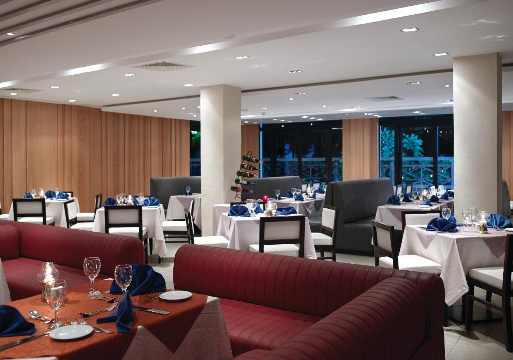 Mūsdienīga restorāna interjers ar elegantām galdiem, baltiem galdautiem un zilo galda piederumu komplektu. Telpa ir apgaismota ar mīkstu gaismu, radot patīkamu atmosfēru.