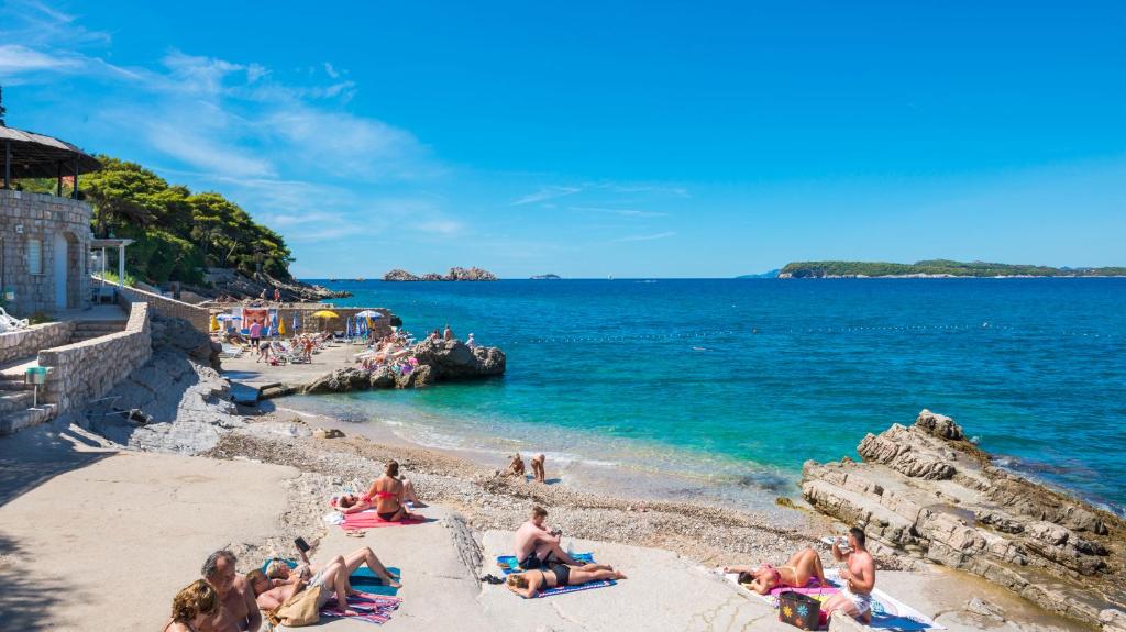 Saulaina pludmale ar cilvēkiem, kas sauļojas un atpūšas pie skaidra ūdens un akmeņainām krastmalām. Fons ar zilo jūru un zaļajiem apkārtnes kokiem.