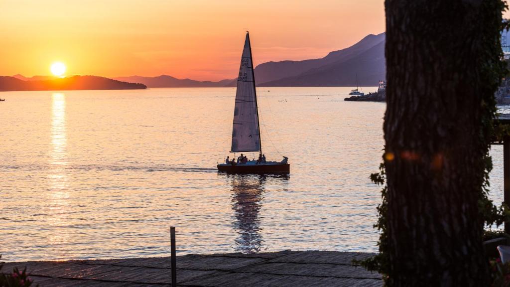 Saulriets virs jūras, kuģis ar burām dodas prom, fons ar kalnu siluetu un mierīgu ūdeni.