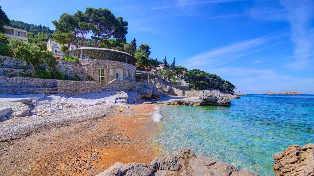 Skaists pludmales skats ar skaidru zilo ūdeni, akmeņiem un zaļiem kokiem, kas ieskauj piekrasti. Mājas un infrastruktūra ir redzama kalnu nogāzē, radot mierīgu atmosfēru.