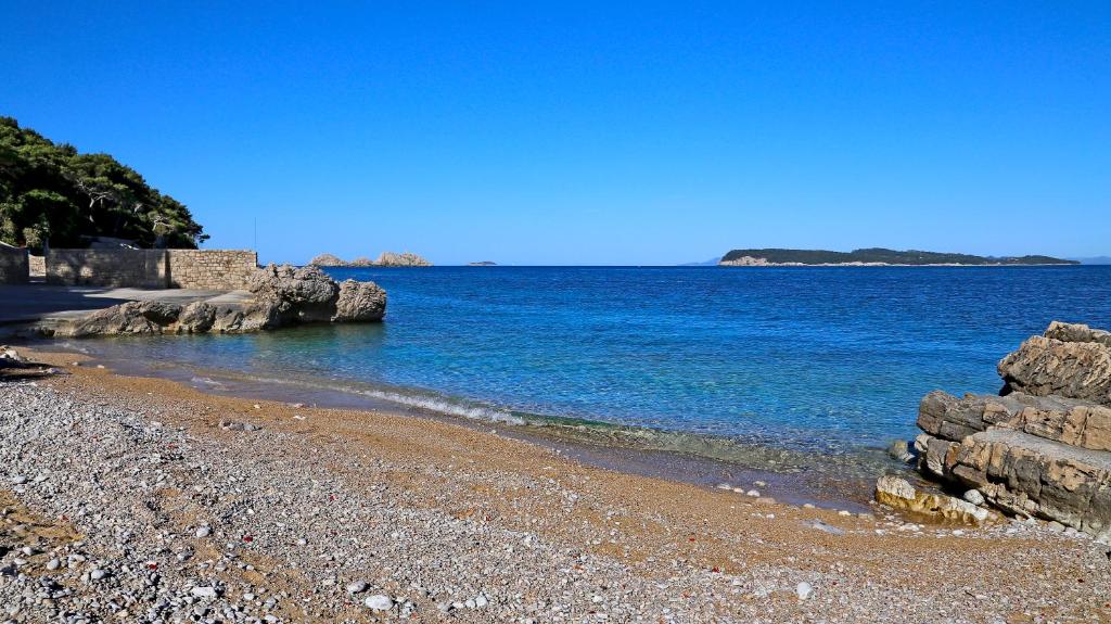 Piekraste ar smilšu pludmali un skaidru zilo ūdeni, kas stiepjas līdz horizonta līnijai, ar akmeņainiem krastiem un apzaļumotu fona.