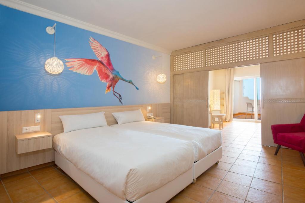 Mūsdienīgs guļamistabas interjers ar divguļamo gultu un krāsainu putna gleznu uz sienas. Telpā ir siltas krāsas apgaismojums un pieejama pieeja uz balkonu.