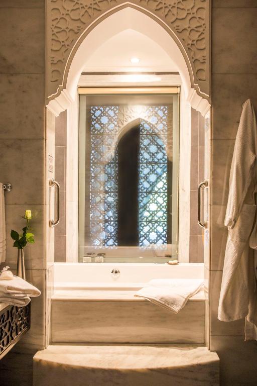 Elegants vannas istabas interjers ar arābu stila elementiem, ietverot marmorētu vannu un dekoratīvu logu ar ornamentiem. Gaisma maigi izgaismo telpu, radot relaksējošu atmosfēru.