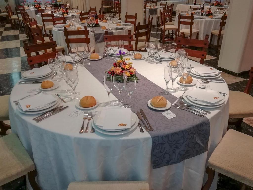 Eleganti noformēts svinību galds ar galda piederumiem un ziedu kompozīciju centrā. Galds ir klāts ar baltu un pelēku galdautu, un uz tā ir maizes rullīši un glāzes.