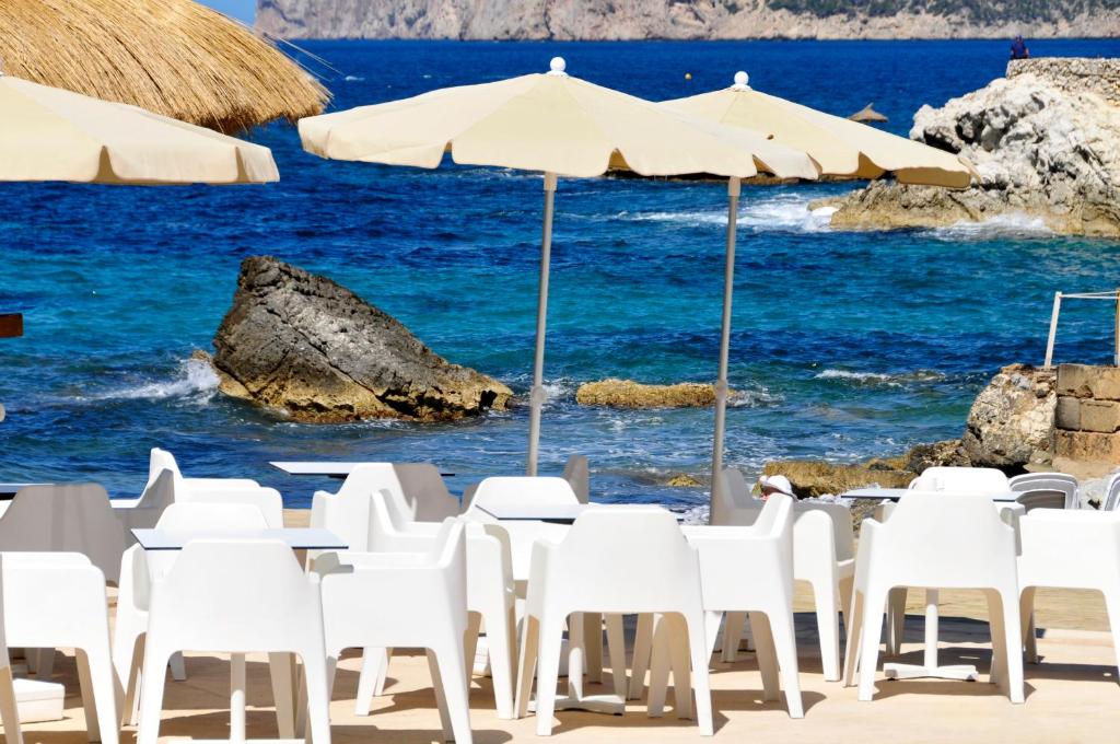 Pludmales kafejnīca ar baltām plastmasas mēbelēm un saulessargiem, blakus skaistai zilai jūrai un akmeņainai krastam.