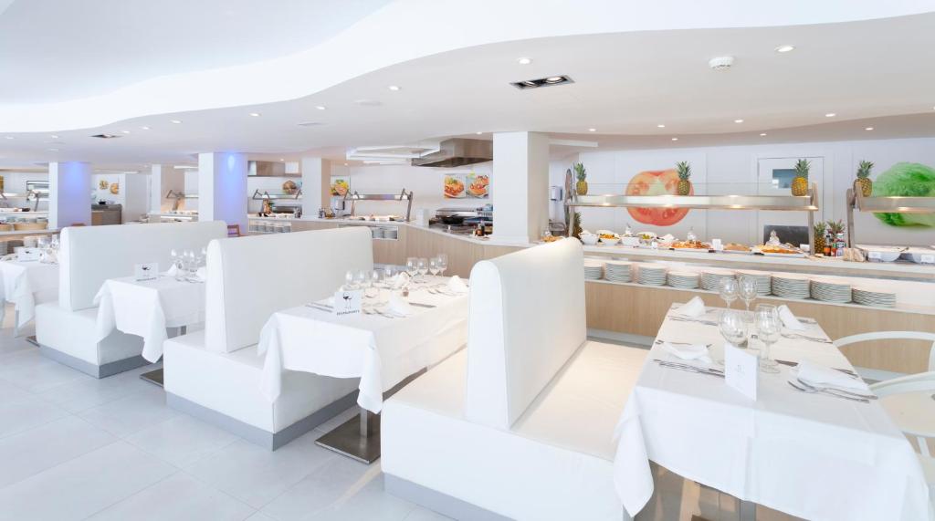 Mūsdienīgs restorāns ar gaišu interjeru, baltām galda segām un komfortablām sēdvietām. Redzama bufete ar dažādiem ēdieniem un augļiem, radot viesmīlīgu atmosfēru.