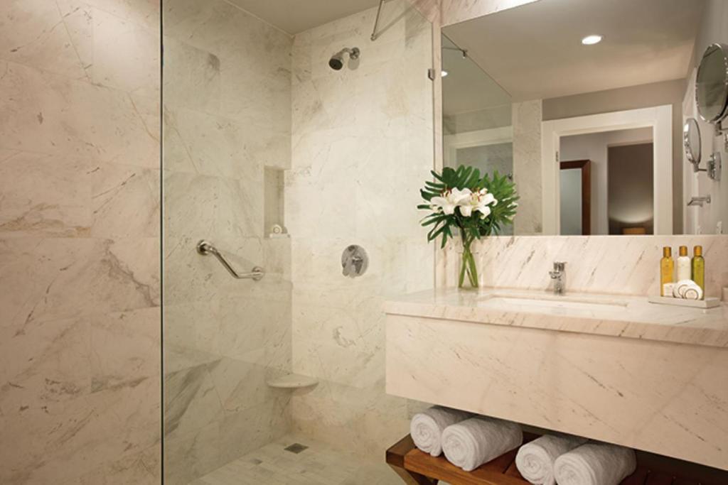 Mūsdienīgs vannas istabas interjers ar marmora apdari, stikla dušu un elegantu izlietni, papildināts ar ziediem un higiēnas precēm.