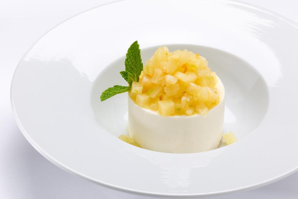 Krēmveida deserts ar augļu gabaliņiem un piparmētru lapu, pasniegts elegantā balta traukā.