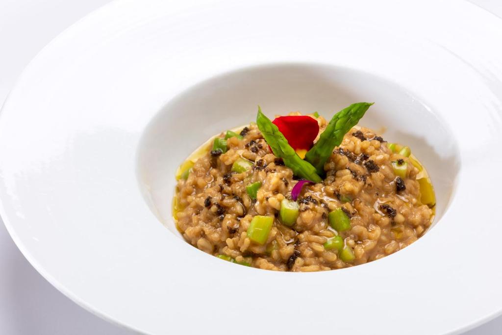 Kremveida risotto ar zaļajiem sparģeļiem un dekoratīvām ziedu lapām, pasniegts eleganta baltā traukā.