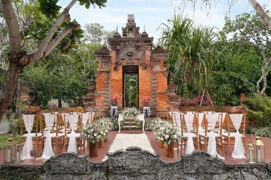 Romantiska kāzu ceremonijas vieta ar tradicionālo Bali arhitektūru, ziedu dekorācijām un koka krēsliem, ieskauta zaļā ainavā.