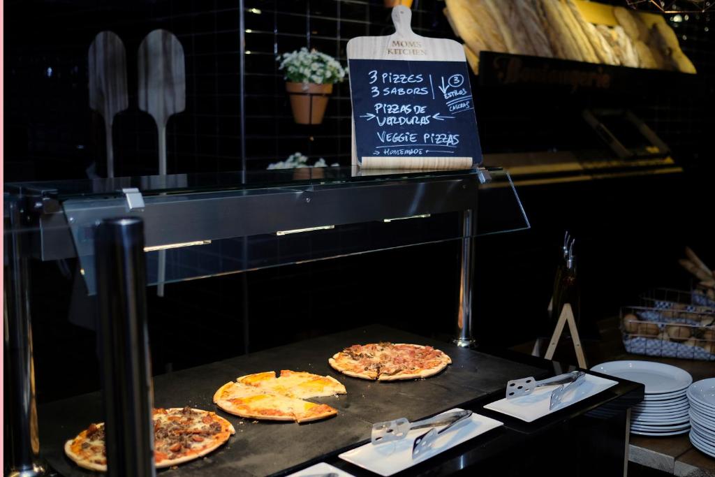 Pica stends ar trīs dažādām picām, ieskaitot dārzeņu variantus, un melnu tāfeli ar piedāvājumiem. Fons ir elegantā restorāna interjers ar maizi un aksesuāriem.