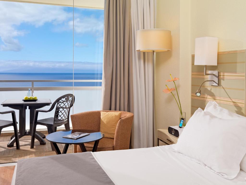 Mūsdienīga viesnīcas numura interjers ar skatu uz jūru, iekārtots ar ērtu gultu, krēslu un galdiņu uz balkona. Svaigi augļi un dekoratīvas puķes papildina relaksējošo atmosfēru.
