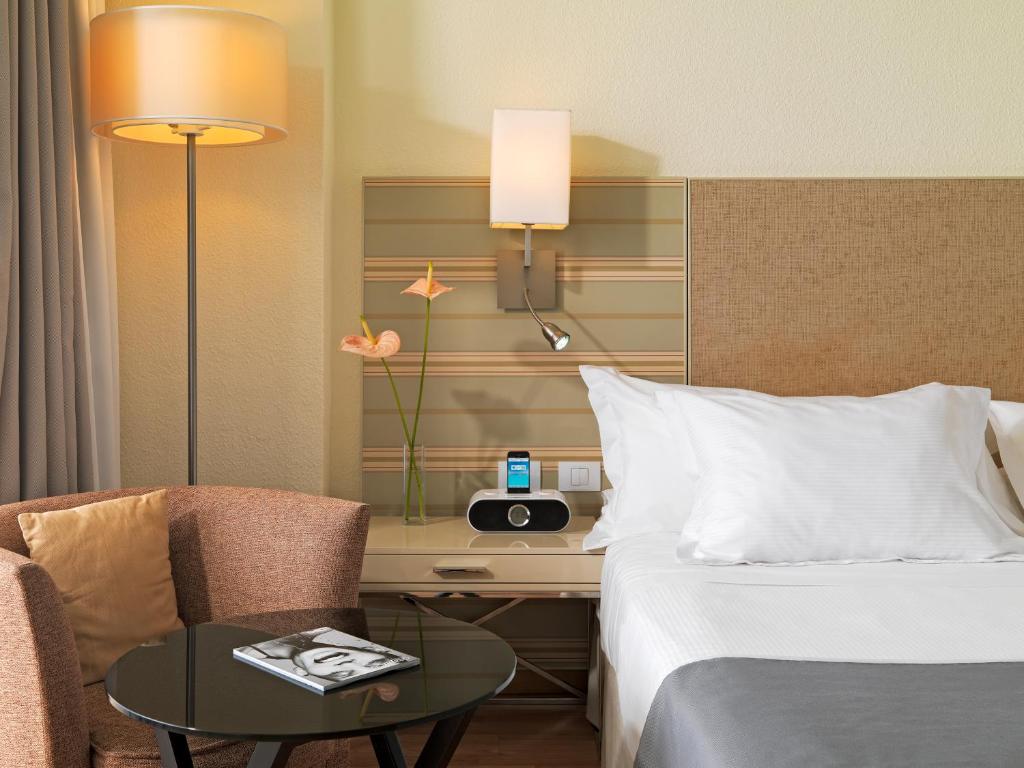 Mūsdienīgs viesnīcas numurs ar ērtu gultu, apgaismojumu un atpūtas stūri, kas ietver krēslus, galdu un dekoratīvas ziedu kompozīcijas.