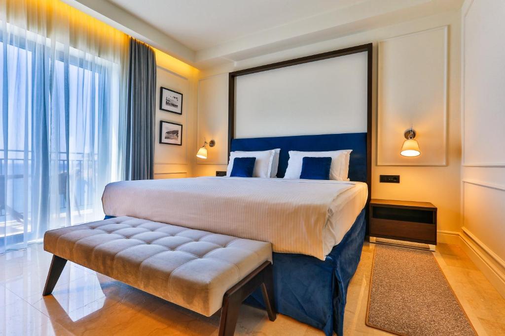 Mūsdienīgs guļamistabas interjers ar plašu gultu, spilveniem un elegantu krēslu. Lielie logi nodrošina dabisko apgaismojumu un skatu uz āru.