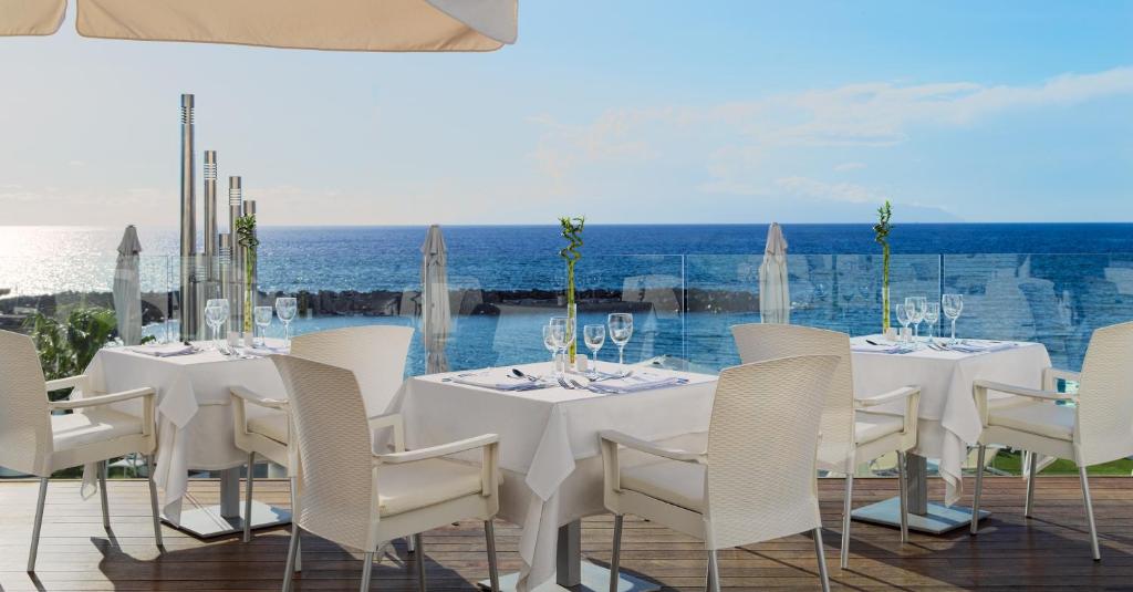 Elegants restorāns ar skatu uz jūru, kurā ir balti galdi un krēsli, kas izvietoti terasē. Fons ietver saulainu debesi un mierīgu ūdeni.