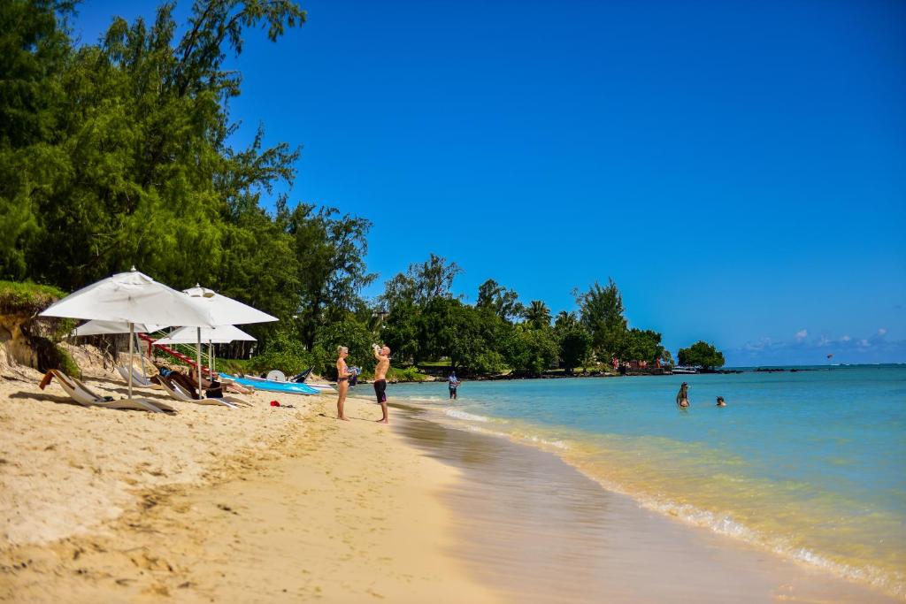 Pludmales ainava ar smiltīm, saulessargiem un cilvēkiem, kas atpūšas un peldas, zem skaidra zila debesu fona.