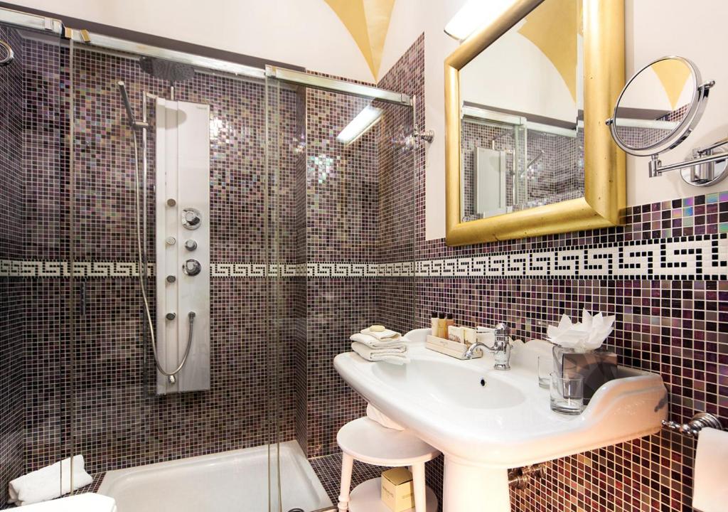 Mūsdienīgs vannasistabas interjers ar stikla dušu un mozaīkas flīzēm, kas izceļ elegantu dizainu un komfortu. Vannasistabā redzams arī liels spogulis un higiēnas piederumi.