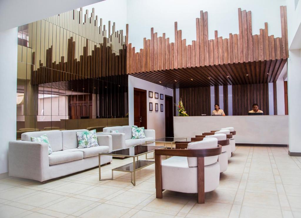 Mūsdienīgs viesnīcas reģistratūras interjers ar elegantiem dīvāniem, stikla galdu un koka akcentiem. Apsveikuma darbinieki stāv pie reģistratūras letes, bet telpā valda gaiša un viesmīlīga atmosfēra.