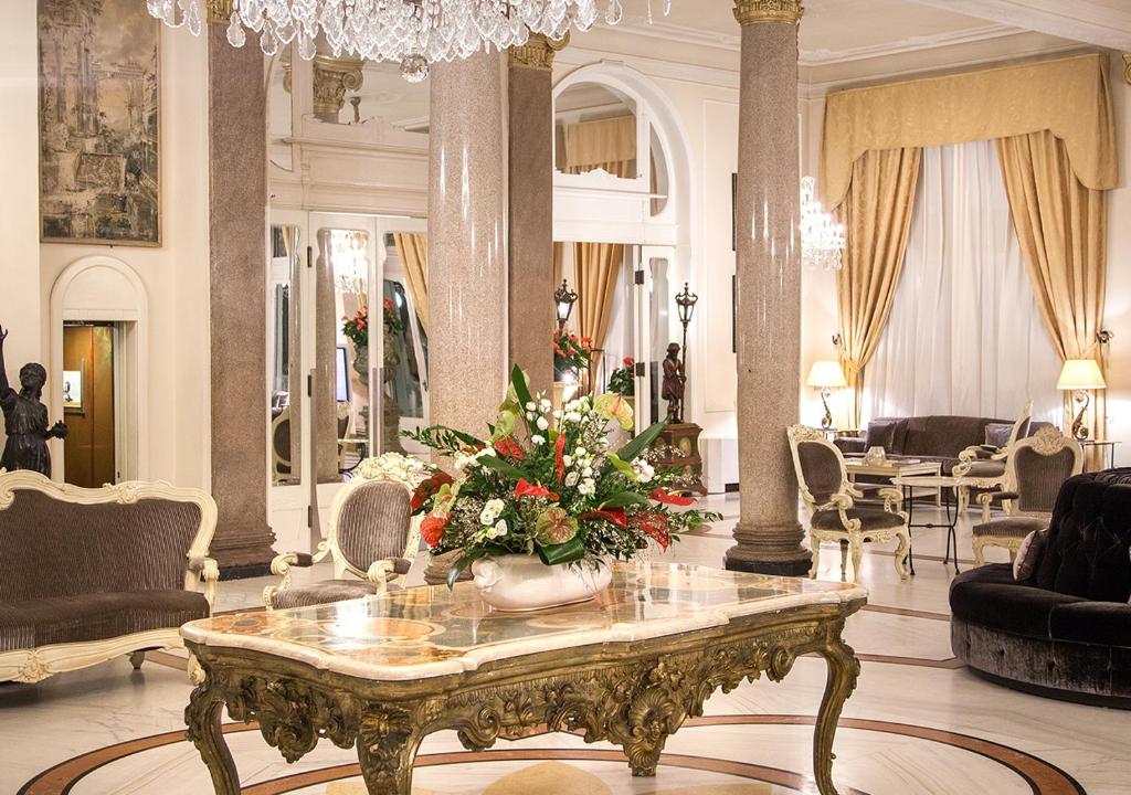 Elegantā viesnīcas halle ar izsmalcinātu interjeru, greznu kristāla lustru un bagātīgu ziedu kompozīciju uz marmora galda. Interjers apvieno klasiskus elementus un modernu komfortu.