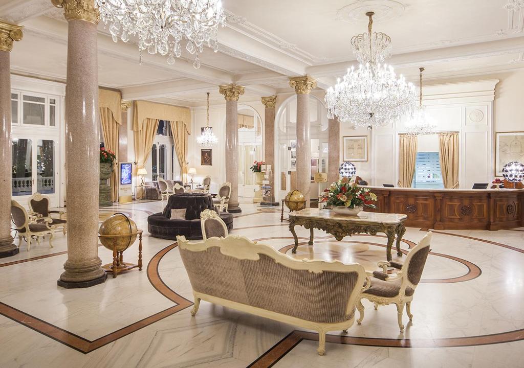 Elegants viesnīcas vestibils ar grezniem lustriem, marmorā veidotu grīdu un stilīgu mēbeļu izkārtojumu. Interjerā ir izsmalcināti elementi un siltas krāsas, kas rada viesmīlīgu atmosfēru.