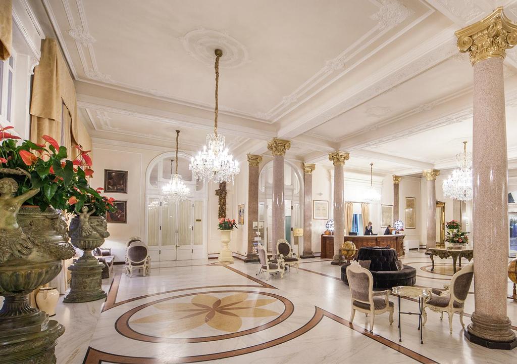 Eleganta viesnīcas vestibila iekšskats ar greznām lustrām, kolonnām un dekoratīvām puķēm. Šis telpas dizains izstaro luksusu un stilu.