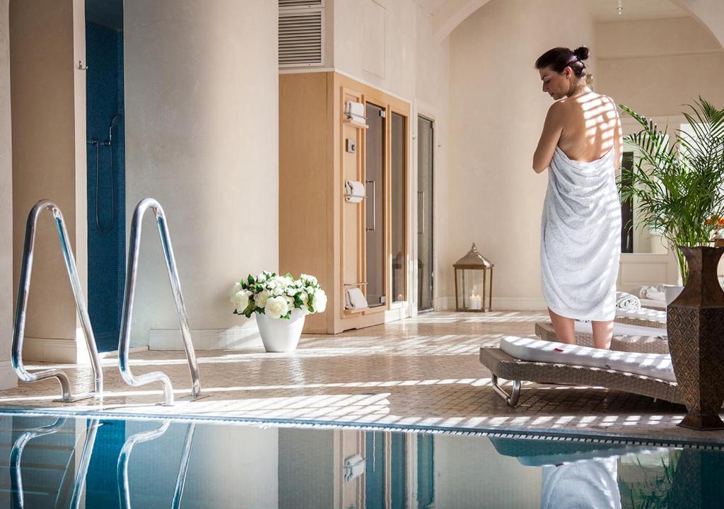 Relaksējošs spa vidus ar sievieti dvielī, kas stāv pie peldbaseina. Telpā ir gaiša apgaismojuma, zaļās augu un ziedu kompozīcijas elementi.