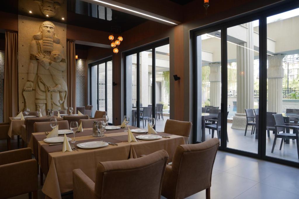 Elegants restorāna interjers ar ērtām krēslām un galdiem, rotāts ar monumentālu skulptūru un plašiem logiem, kas ved uz āra terasi.