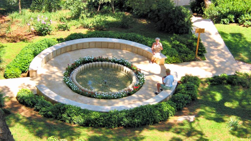 Dārzs ar apaļu strūklaku centrā, ap to izvietoti ziedi un zaļumi. Divi cilvēki atpūšas pie strūklakas, baudot mierīgu atmosfēru.