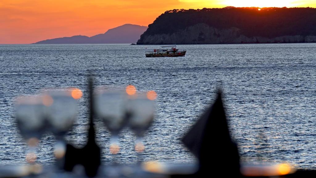 Saulrieta skats uz jūru ar zvejas laivu un vīna glāzēm priekšplānā. Siltas krāsas piepilda debesus, radot romantisku atmosfēru.