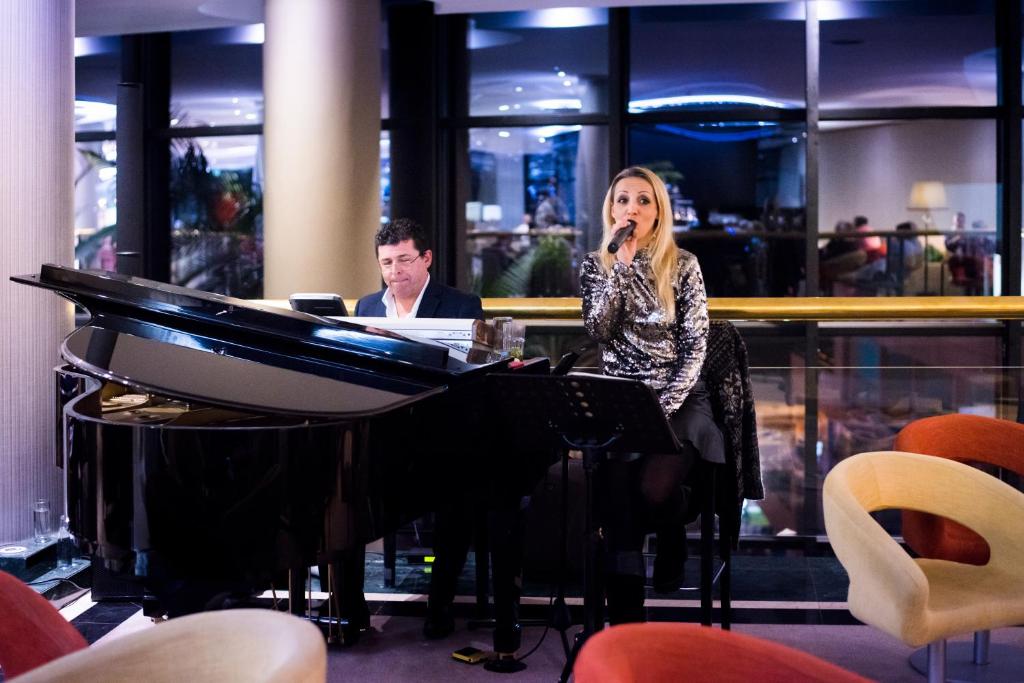 Mūziķi uzstājās luksusa viesnīcas bārā, kur pianists spēlē pie klavierēm, bet dziedātāja izpilda melodiju. Aiz viņiem redzama eleganta viesnīcas vide ar gaismas efektiem.