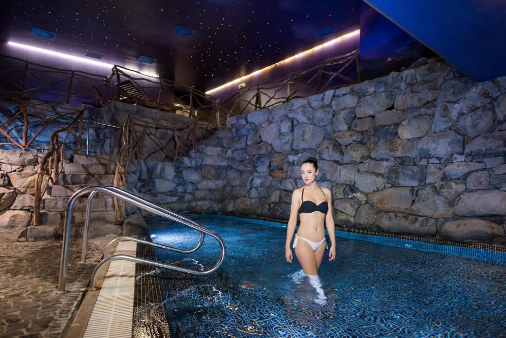 Sieviete staigā pa baseinu, kas atrodas eleganti iekārtotā spa vidē ar akmens sienām un zvaigžņotu griestu apgaismojumu.
