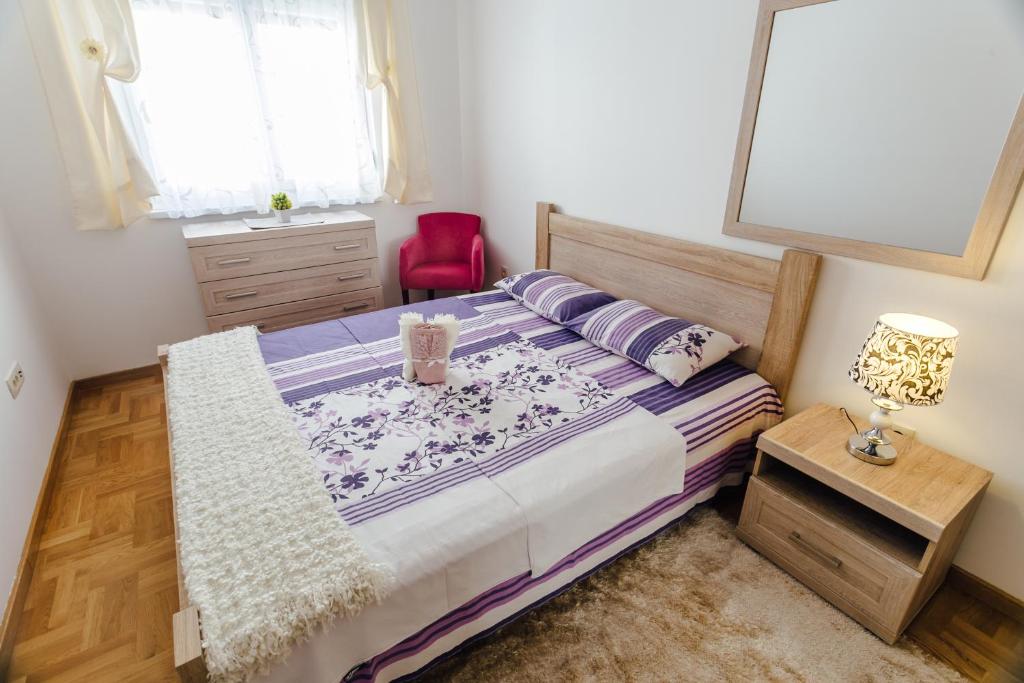 Mūsdienīga guļamistaba ar gultu, kas pārklāta ar violetu un baltu gultas veļu, dekoratīvu lampu un sarkanu krēslu. Telpa ir gaiša un mājīga, ar koka grīdām un logu ar gaisīgu aizkaru.