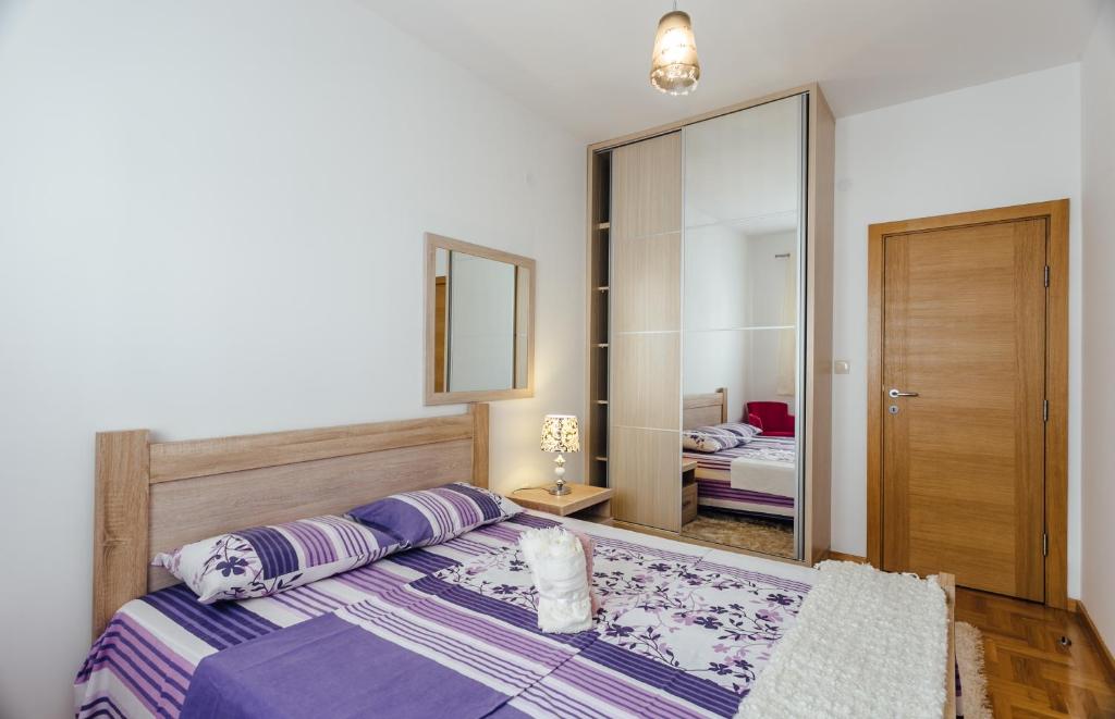 Mūsdienīga guļamistaba ar koka gultu un spoguļa skapī, rotāta ar violetiem un ziedu rakstiem. Siltas krāsas un eleganta apgaismojuma elementi rada mājīgu atmosfēru.
